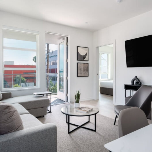 Modern living room with gray sofas, a round coffee table, wall-mounted TV, and minimalist decor. Large windows and an open door let in natural light; a bedroom is visible in the background.