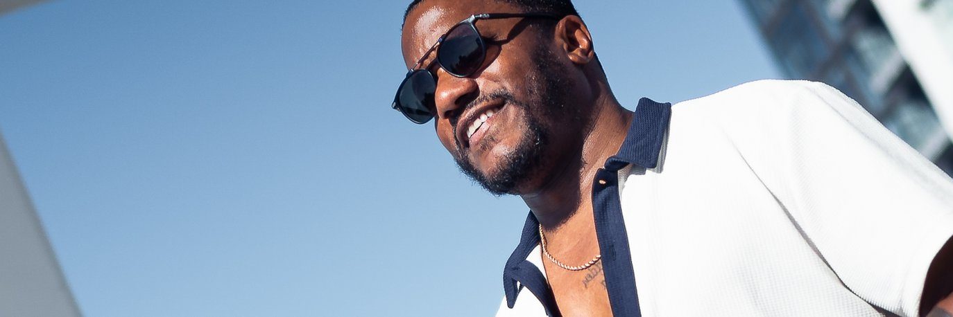 A man wearing sunglasses and a white shirt with a dark collar smiles outdoors on a sunny day, with a blue sky and a modern building in the background.