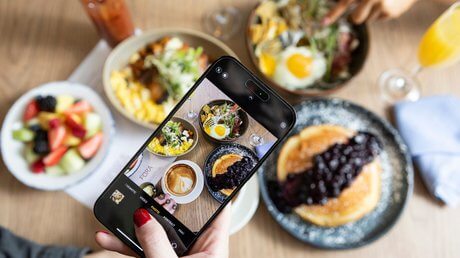 A person takes a photo of a brunch table with their phone, capturing plates of pancakes with berry sauce, a fruit bowl, eggs, and coffee. Another hand reaches toward a bowl in the background.