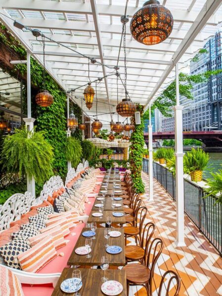 A long outdoor dining area features wooden tables, colorful plates, patterned cushions, hanging lanterns, and lush greenery, set alongside a river with city buildings in the background.
