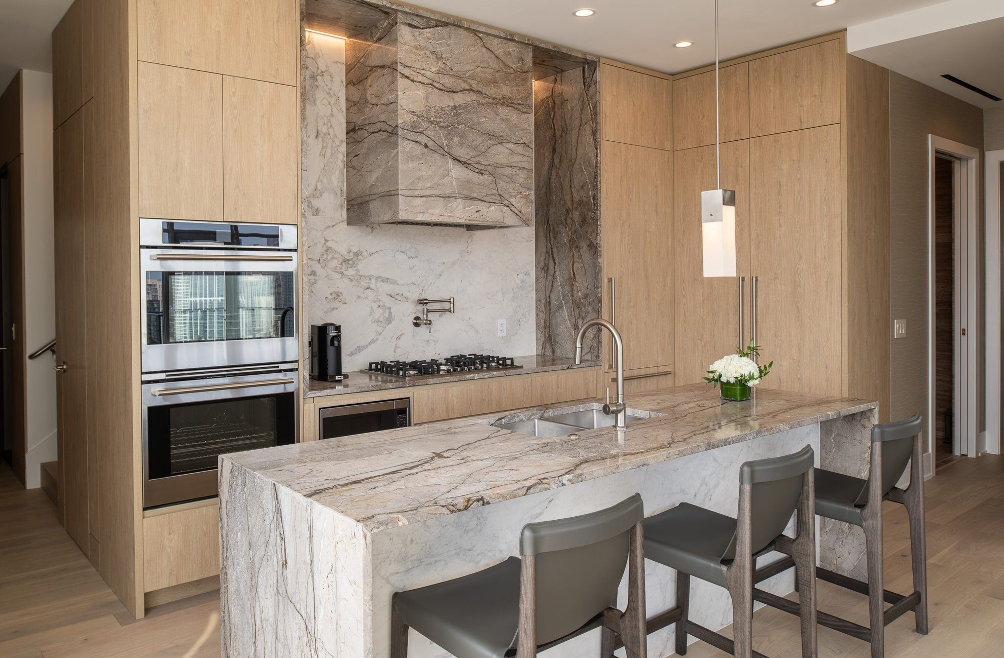 Modern kitchen with light wood cabinets, marble countertops and backsplash, built-in oven, gas cooktop, stainless steel fixtures, and three gray bar stools at an island. A vase with white flowers sits on the island.