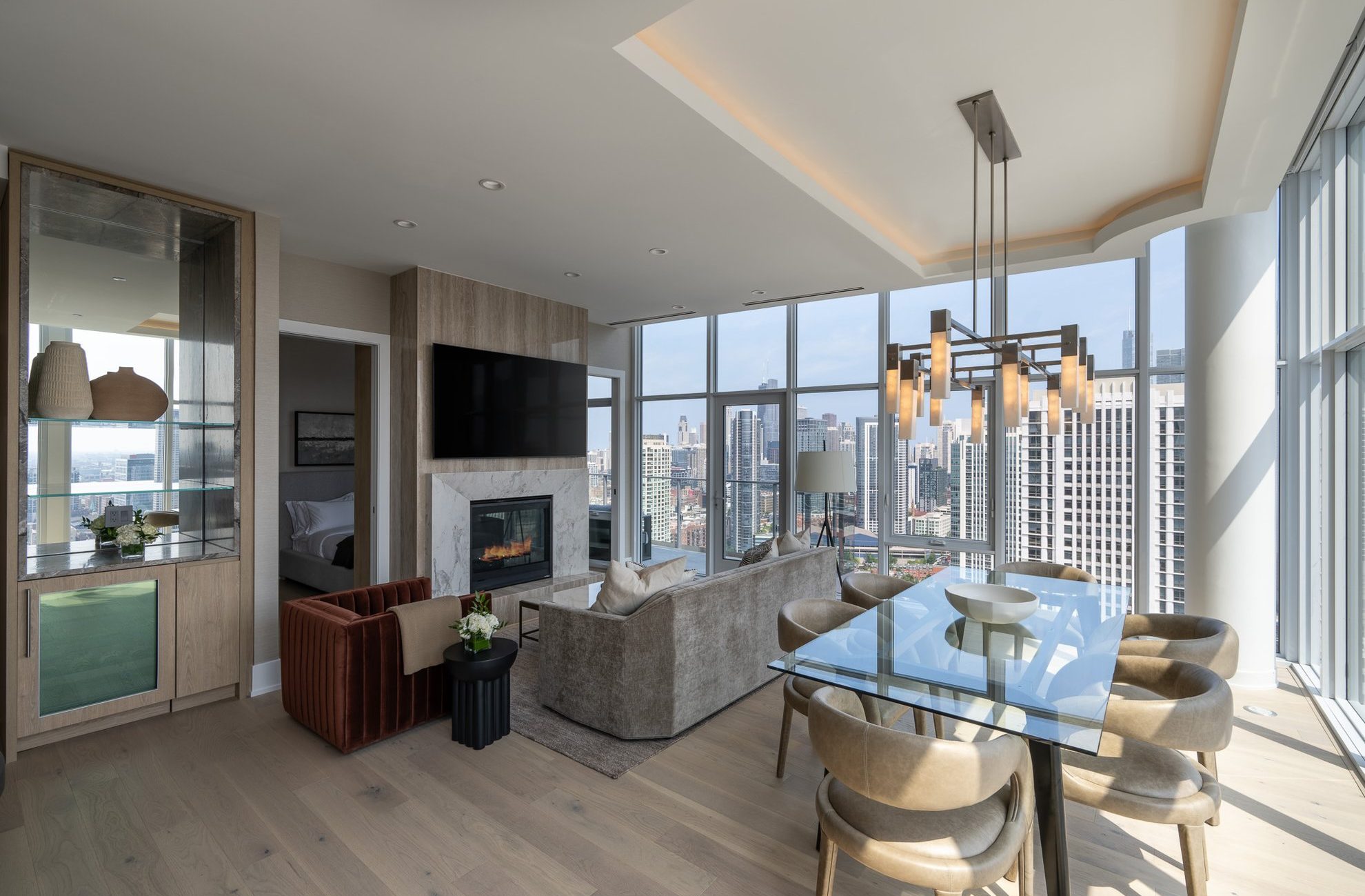 Modern high-rise apartment with floor-to-ceiling windows, a city view, neutral-toned furniture, a fireplace, wall-mounted TV, and a glass dining table with six chairs under a contemporary chandelier.