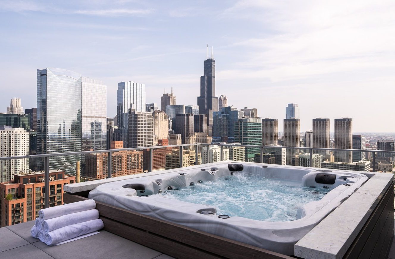 A rooftop hot tub with folded white towels on its edge overlooks a modern city skyline with tall skyscrapers under a clear sky.