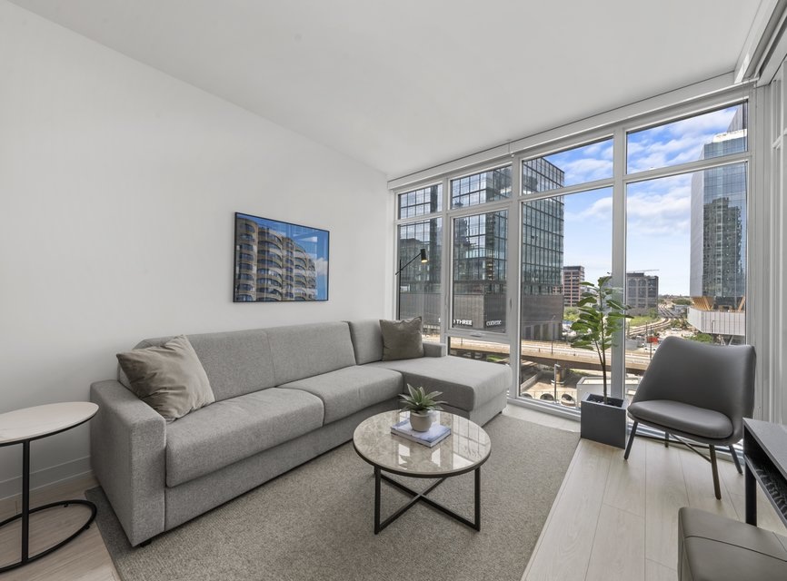 Modern living room with a gray sectional sofa, round coffee table, single gray chair, and large floor-to-ceiling windows overlooking city buildings and streets on a sunny day.