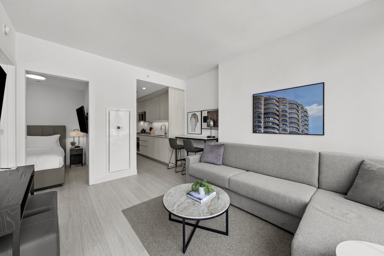 Modern open-plan living space with a gray sectional sofa, round coffee table, wall art, compact kitchen with bar stools, and a bedroom visible through an open doorway. Decor is minimalist with neutral tones.