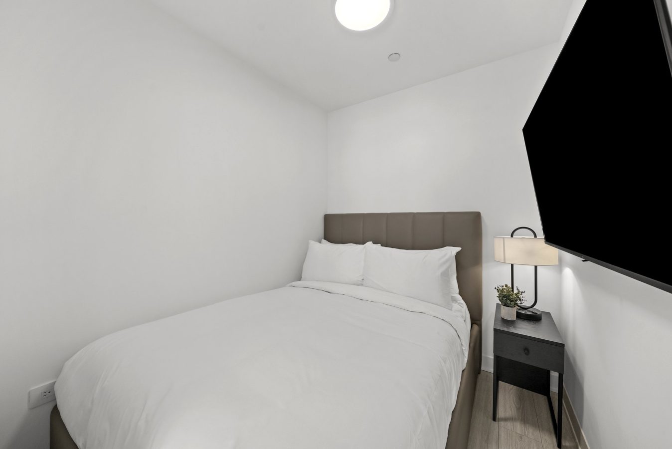 A small, modern bedroom with white walls, a neatly made bed with white bedding, a gray headboard, a black nightstand with a lamp and plant, and a large wall-mounted TV.