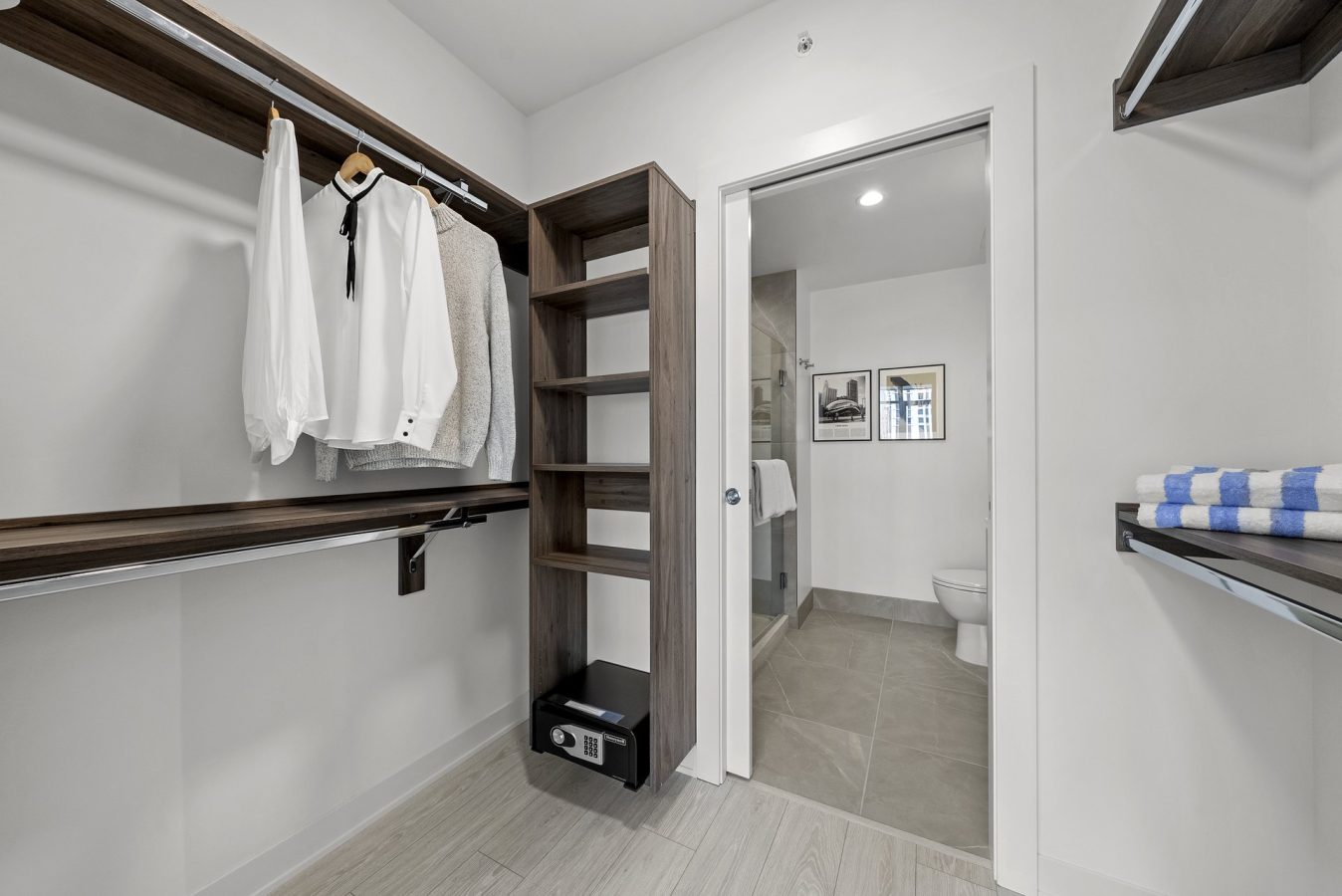 A modern walk-in closet with wooden shelves, hanging clothes, a small safe, and folded towels, leading to a bathroom with tile flooring and framed pictures on the wall.