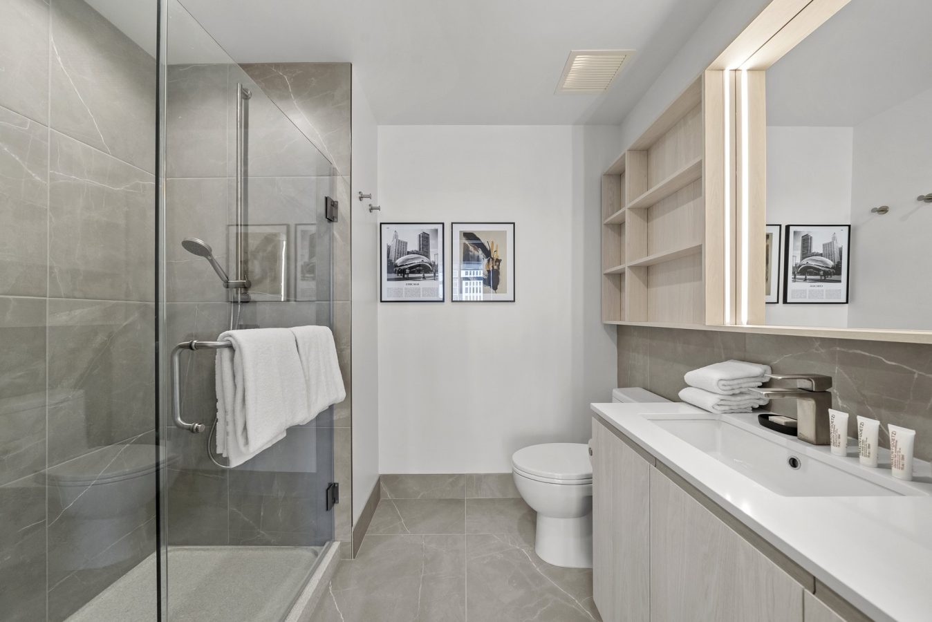 Modern bathroom with a glass shower, gray tile walls and floor, a white toilet, a wide sink with a light wood vanity, open shelves, a large mirror, and two framed pictures on the wall. Towels and toiletries are neatly arranged.