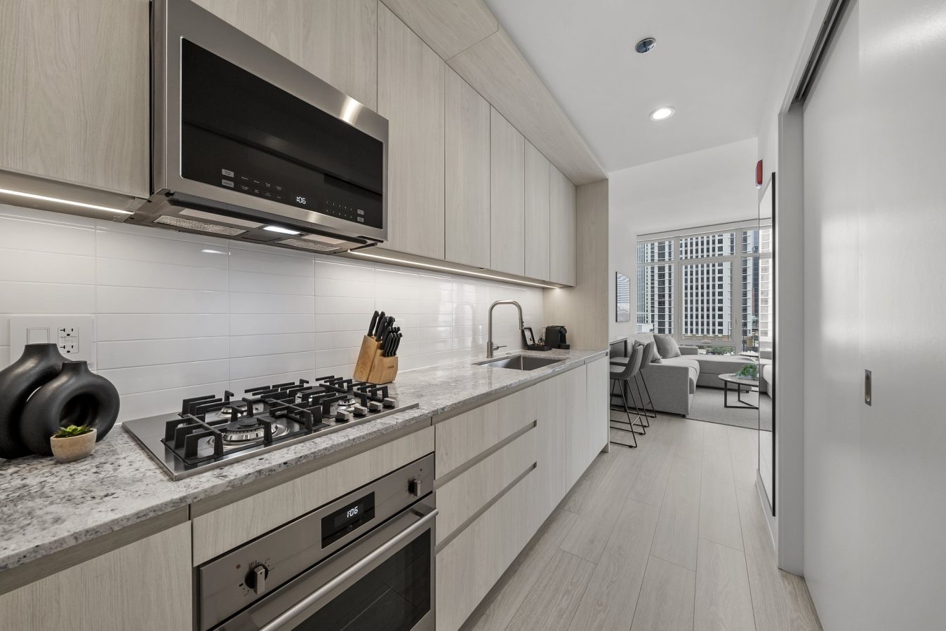 Modern galley kitchen with light wood cabinets, built-in oven, gas stovetop, and marble countertop; open view to a bright living area with large windows and cityscape in the background.