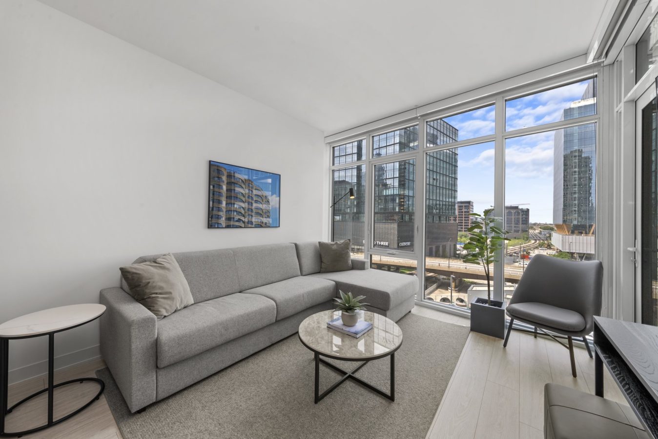 Modern living room with a gray sectional sofa, round coffee table, side table, and large windows offering a view of city buildings and blue sky. A potted plant and wall art complete the minimalist decor.