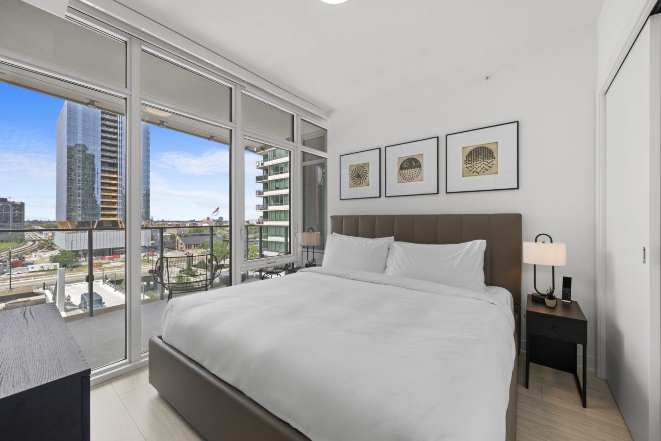 Modern bedroom with large windows, a king-sized bed with white bedding, two nightstands, and framed art on the wall. The windows overlook a cityscape with tall buildings and a blue sky.