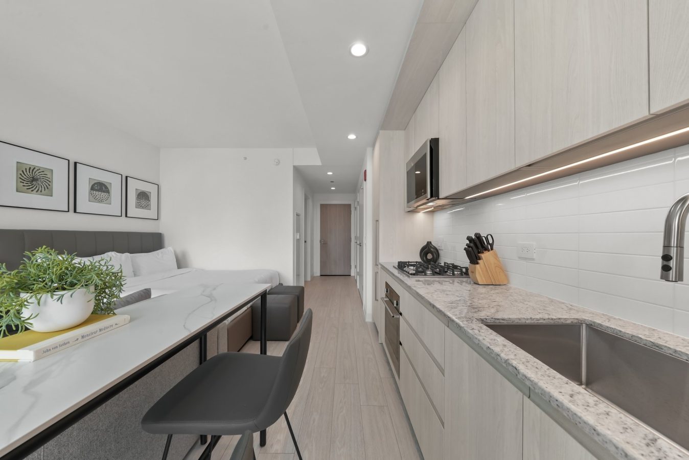 Modern studio apartment with a sleek kitchen, light wood cabinets, marble countertop, built-in stove, and sink; bed and framed art are visible in the background along with a small dining area and plant.