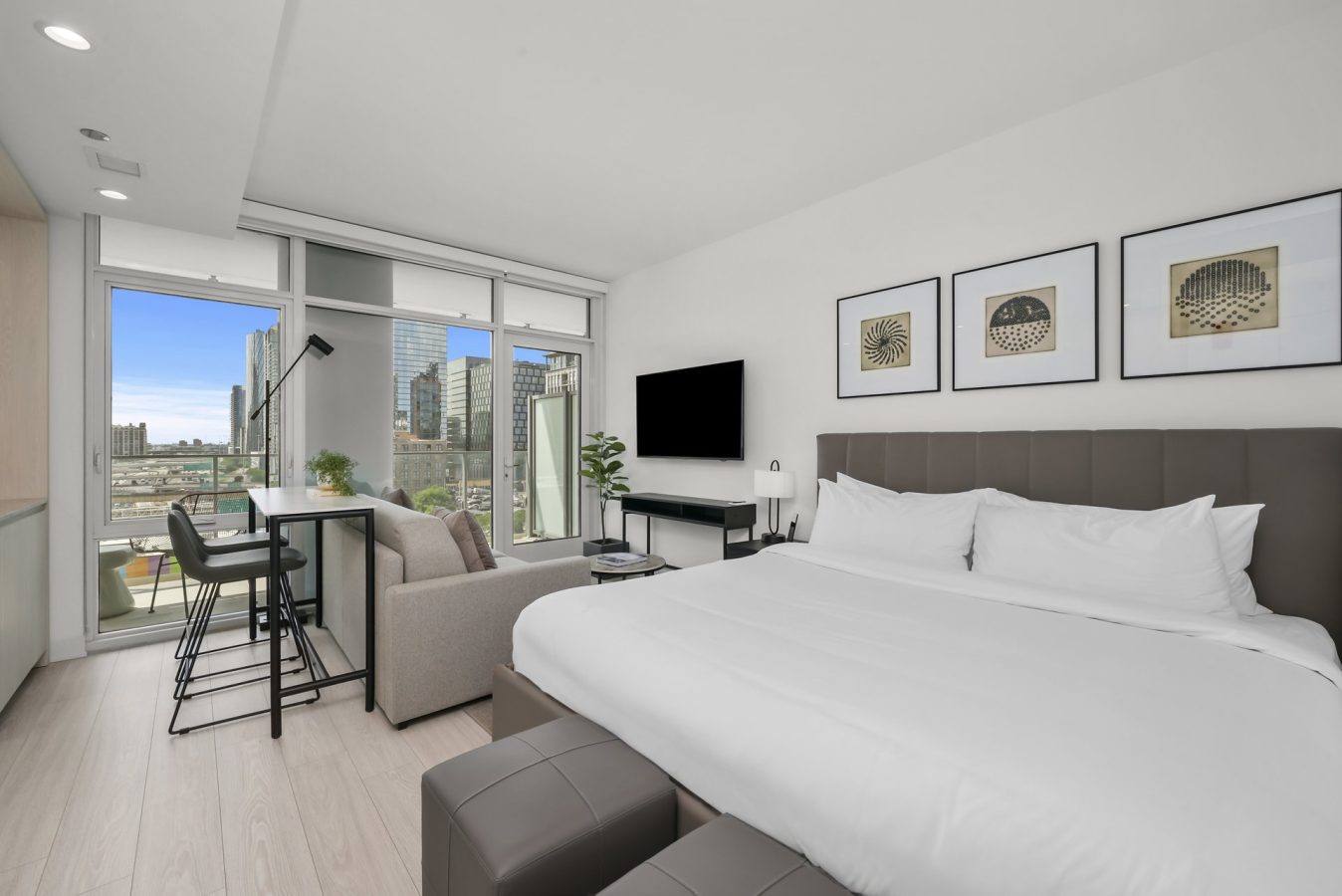 A modern studio apartment with a large bed, sofa, wall-mounted TV, dining table, and floor-to-ceiling windows offering a city view. Minimalist decor includes framed art and a potted plant.