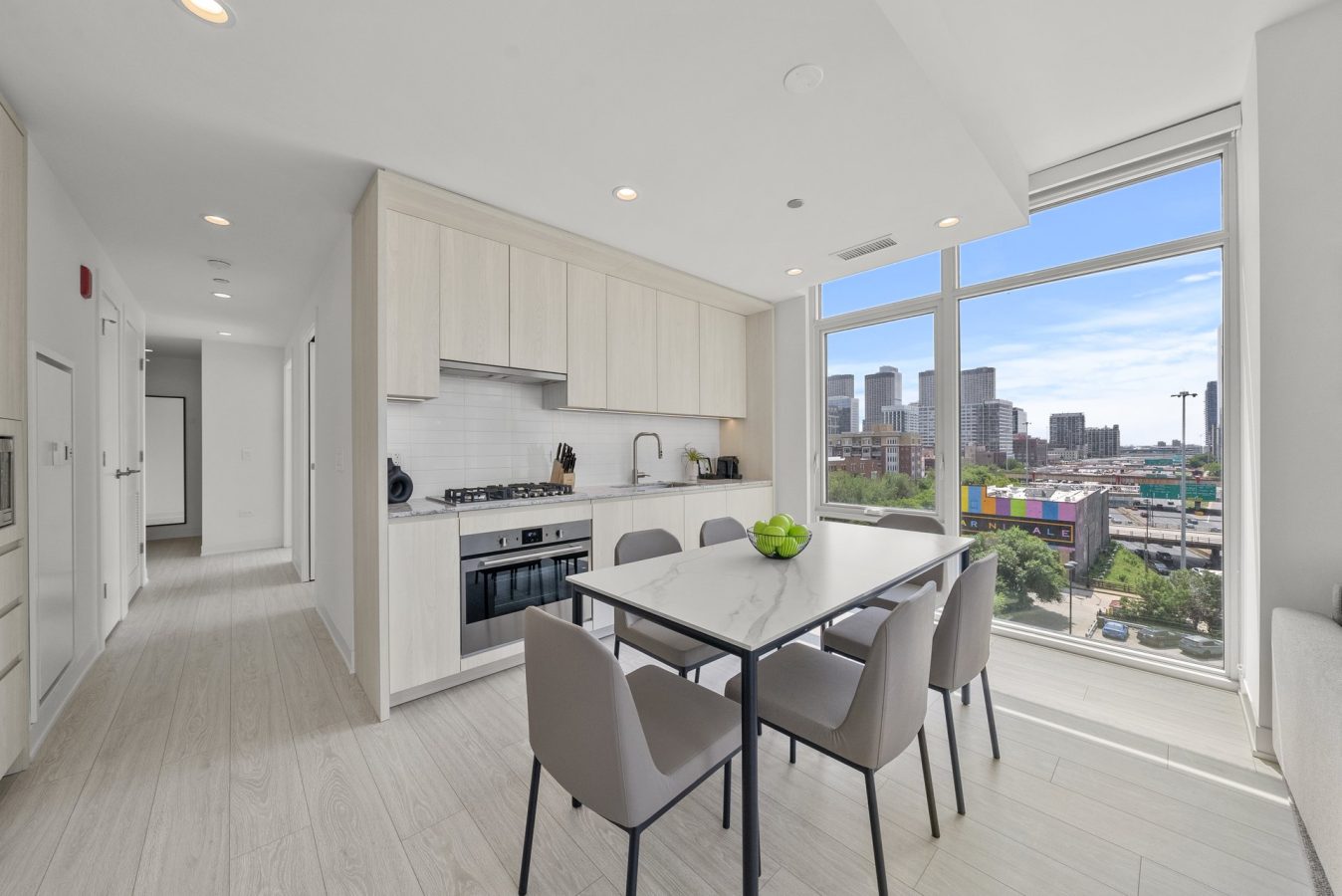 A modern kitchen and dining area with light wood cabinets, a built-in oven, a white table with four gray chairs, and large windows offering a cityscape view. Natural light fills the bright, minimalist space.