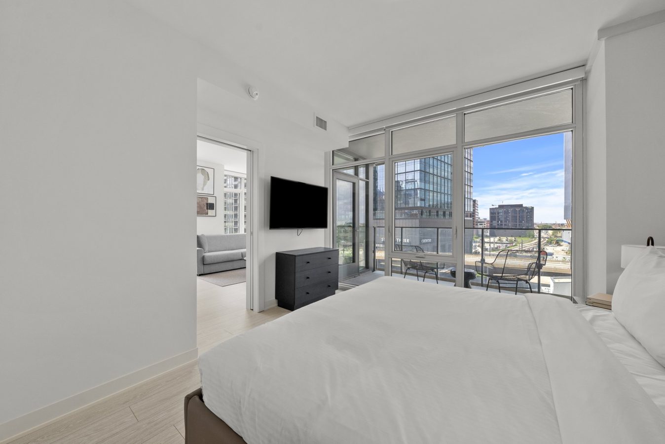 Modern bedroom with a large bed, wall-mounted TV, dresser, and floor-to-ceiling windows leading to a balcony with seating. Cityscape views and an adjoining room with a sofa are visible.