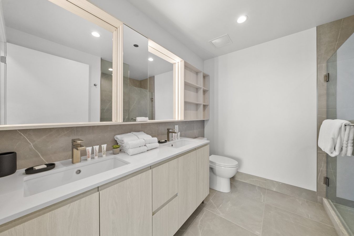 Modern bathroom with light wood cabinets, a wide mirror, neatly folded towels, toiletries on the counter, a toilet, built-in shelves, and a glass-enclosed shower with a towel hanging inside. Neutral beige and white color scheme.