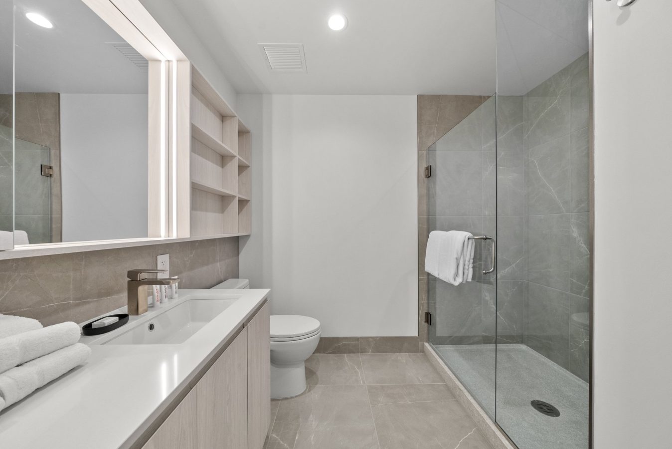 Modern bathroom with a large mirror, sink with towels and toiletries, a toilet, built-in shelves, and a glass-enclosed shower with gray tiles and a white towel hanging on the shower door.
