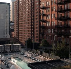 A modern cityscape with tall brick and glass apartment buildings overlooking a rooftop pool area lined with lounge chairs, cabanas, and greenery in the foreground.