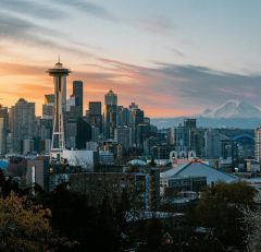 Seattle skyline at sunset with the Space Needle in the foreground, skyscrapers in the background, and Mount Rainier visible in the distance under a partly cloudy sky.