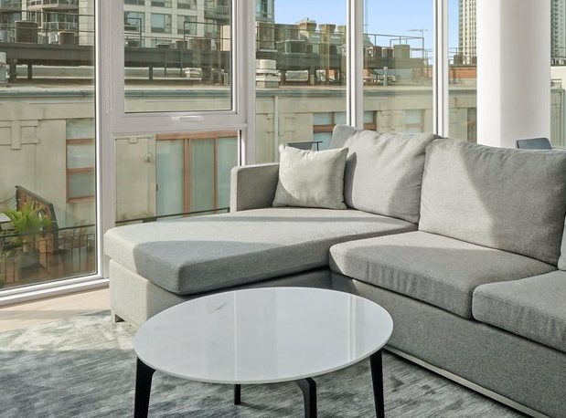 A modern living room with a gray sectional sofa, a round white coffee table, and large windows offering a city view with tall buildings outside.