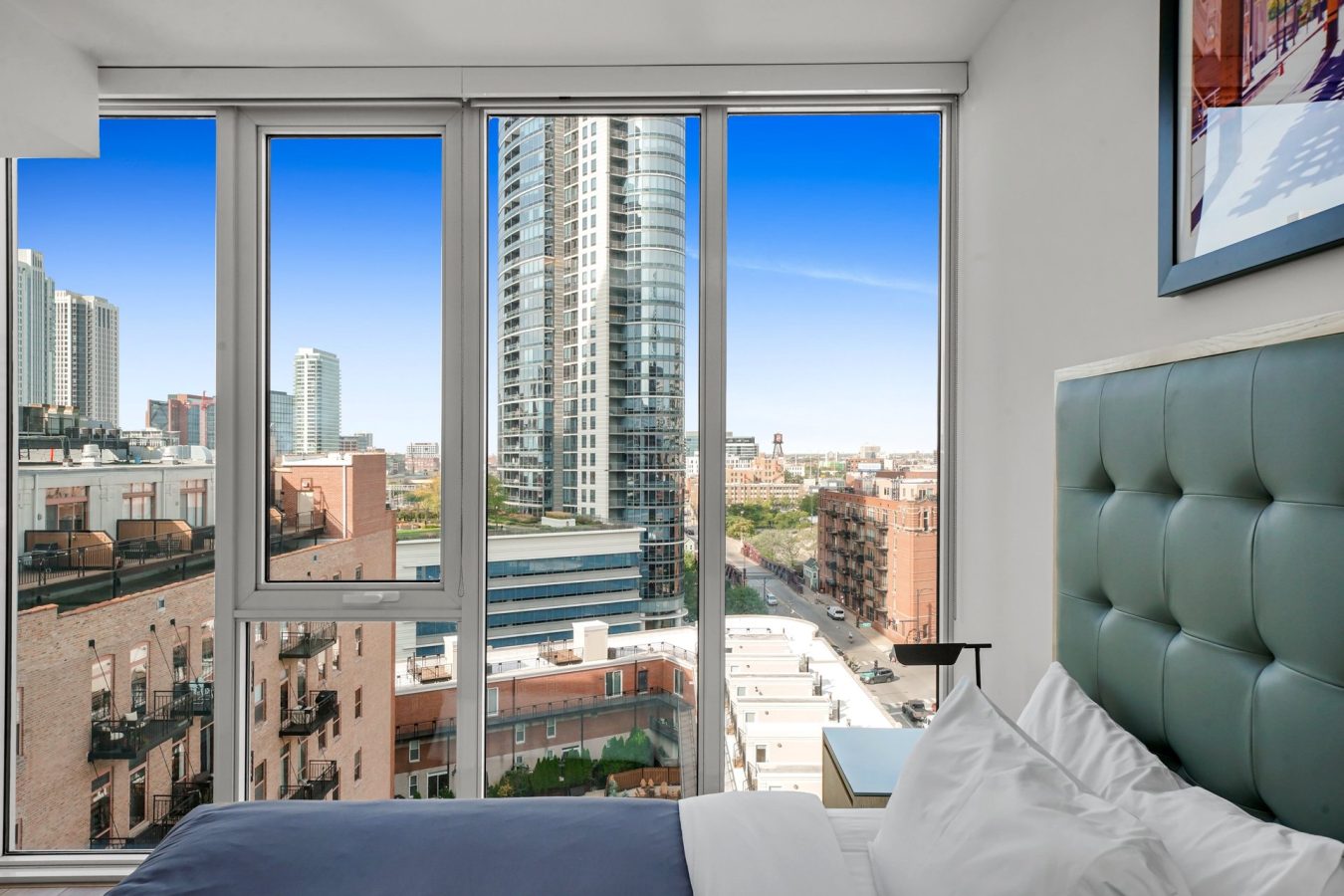 A modern cityscape view through large floor-to-ceiling windows from a bedroom with a neatly made bed and blue headboard, high-rise buildings visible outside under a clear blue sky.
