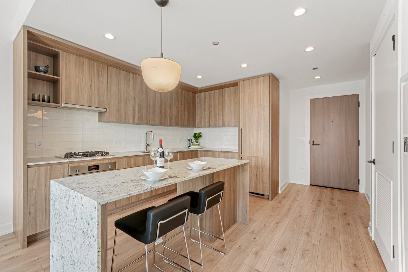 Modern kitchen with light wood cabinets, white tile backsplash, and a marble island with two black bar stools. The space is well-lit with recessed lighting and a hanging pendant lamp above the island.