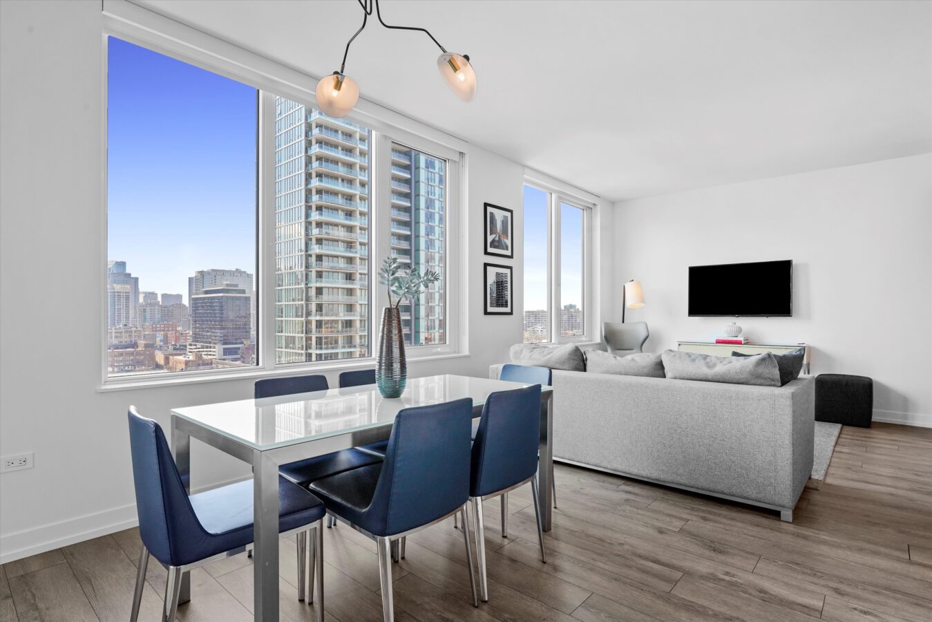 Modern apartment with large windows, a dining table with blue chairs, a gray sofa, wall-mounted TV, and cityscape views from high-rise windows. The space is bright with contemporary decor and wood flooring.