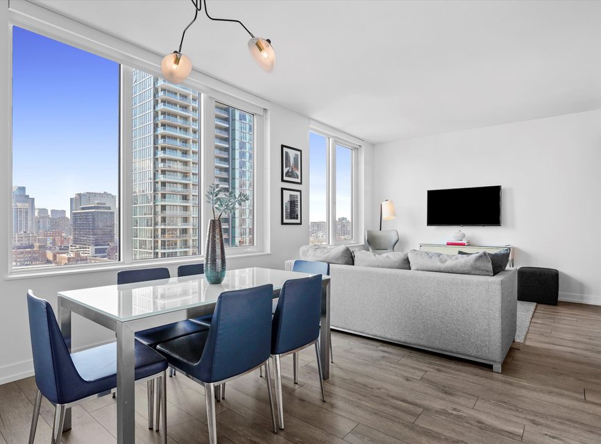 Modern apartment interior with large windows, city views, a dining table with blue chairs, a gray sofa, TV mounted on a white wall, and light wood flooring. Bright and airy with contemporary decor.