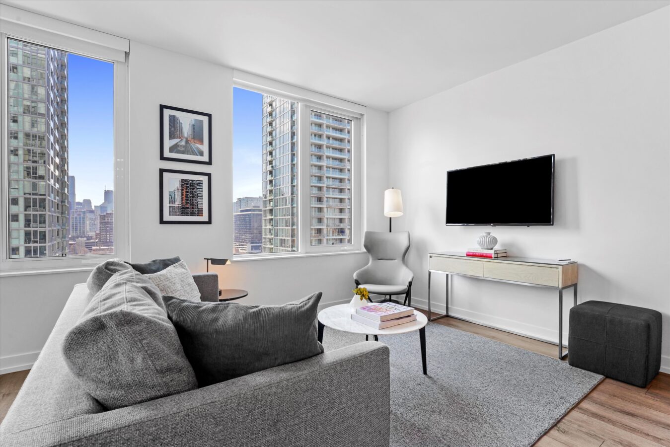 Modern living room with large windows showing city views, a gray sofa, round white coffee table with books, an armchair, TV on a console, floor lamp, and neutral decor. Natural light brightens the space.