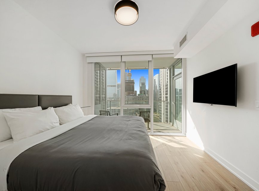 Modern bedroom with a large bed, gray bedding, wall-mounted TV, and floor-to-ceiling windows offering a city skyline view. Light wood flooring and a balcony are visible, creating a bright, airy atmosphere.