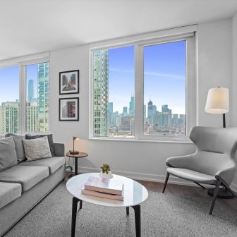 A modern living room with a large window offering a city skyline view. The room features a gray sofa, an armchair, a round coffee table with books, framed wall art, a floor lamp, and neutral decor.