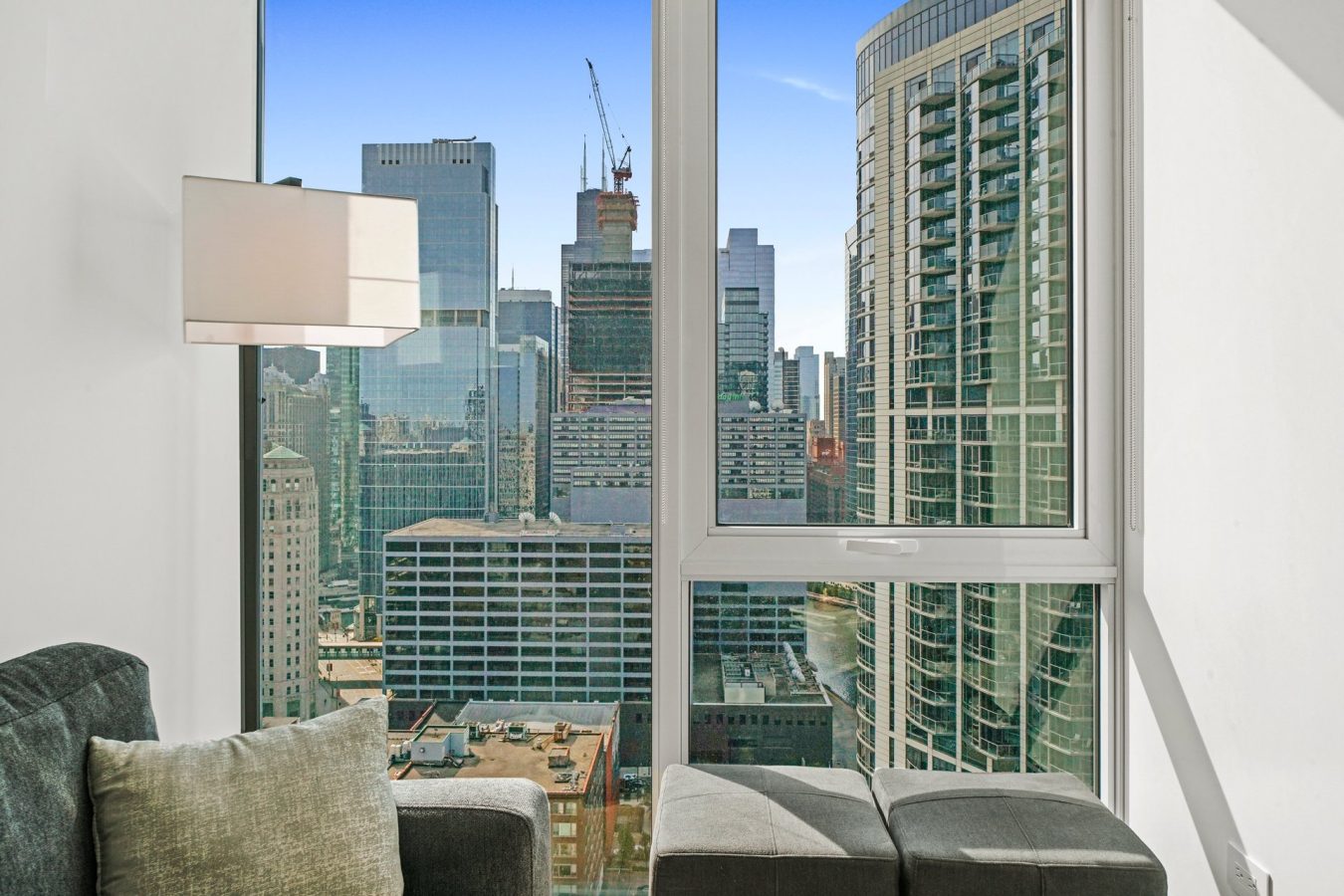 A bright city apartment with a gray sofa and ottoman sits by a large window, offering a clear view of modern high-rise buildings and a blue sky outside. A wall-mounted lamp is visible to the left.