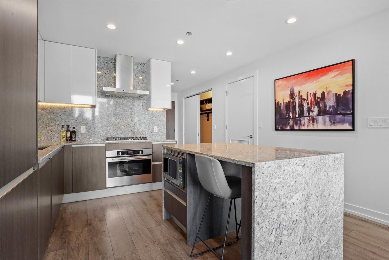 Modern kitchen with wood flooring, sleek cabinets, built-in oven, gas stove, gray stone countertops, a central island with a chair, and a colorful cityscape painting on a white wall.