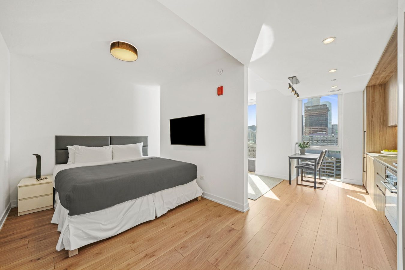 Modern, bright studio apartment with a large bed, mounted TV, wooden floors, small dining area by sunny windows, and a compact kitchen on the right. Cityscape visible through floor-to-ceiling windows.