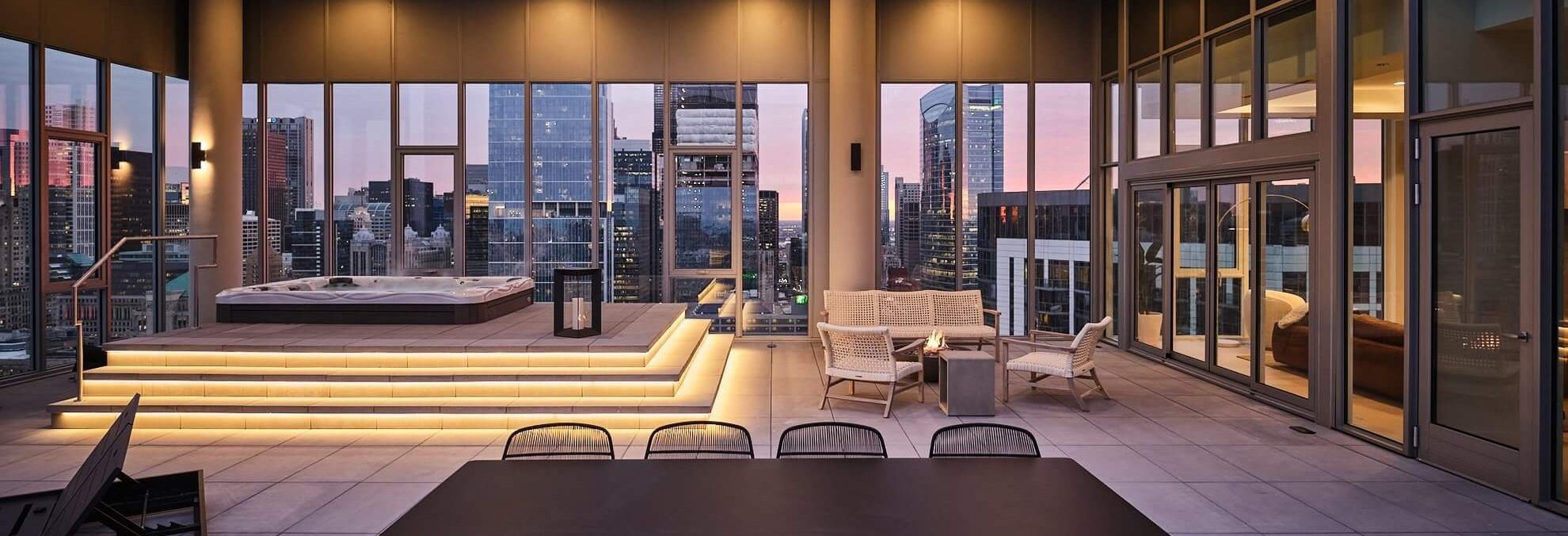 A modern rooftop lounge with large windows overlooking a city skyline at sunset, featuring a raised hot tub, outdoor seating, and sleek furniture on a spacious tiled terrace.