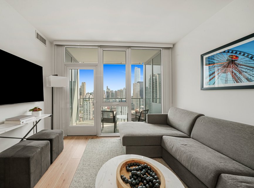 Modern living room with a gray sectional sofa, wall-mounted TV, round coffee table, and large glass doors leading to a balcony overlooking a city skyline. Framed ferris wheel photo hangs on the wall.