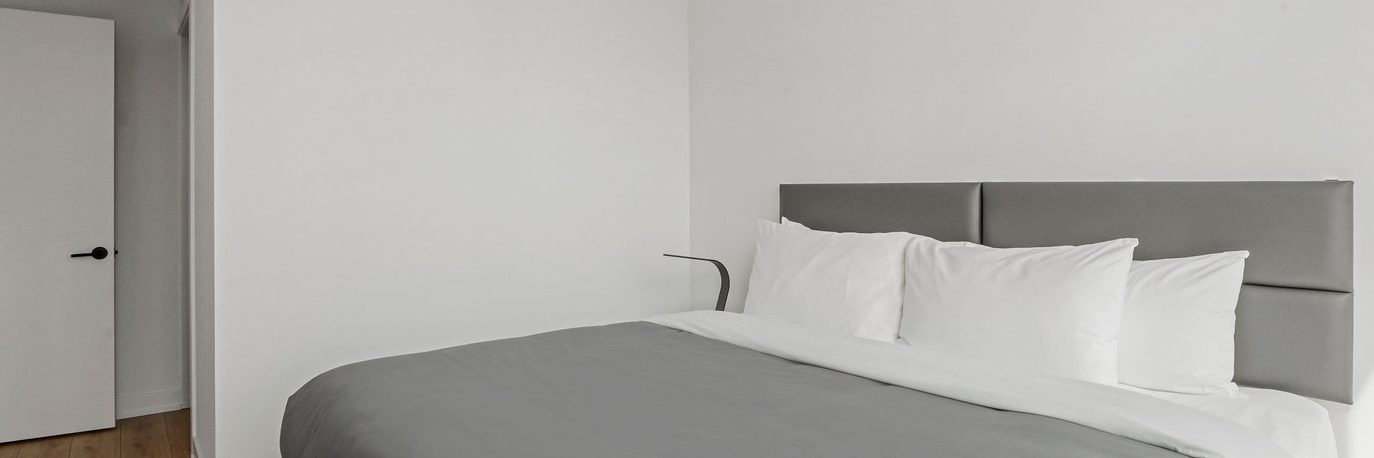 A modern, minimal bedroom with a gray upholstered headboard, white pillows, gray blanket, and white walls. An open door is visible to the left, and the room has a clean, uncluttered look.