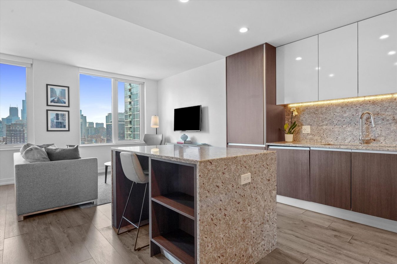 Modern apartment with a kitchen featuring wood and white cabinets, granite countertops, a sink, and open shelves. The living area has a gray sofa, a TV, large windows, and city skyline views.