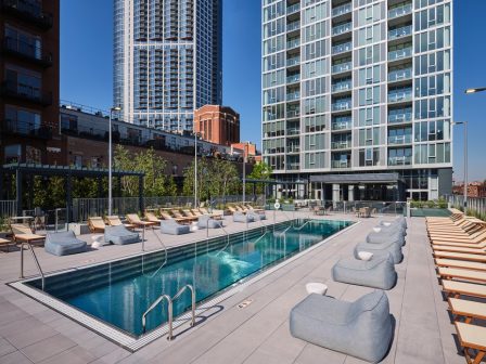 A modern outdoor swimming pool surrounded by lounge chairs and cushioned seating, with tall apartment buildings and a clear blue sky in the background.