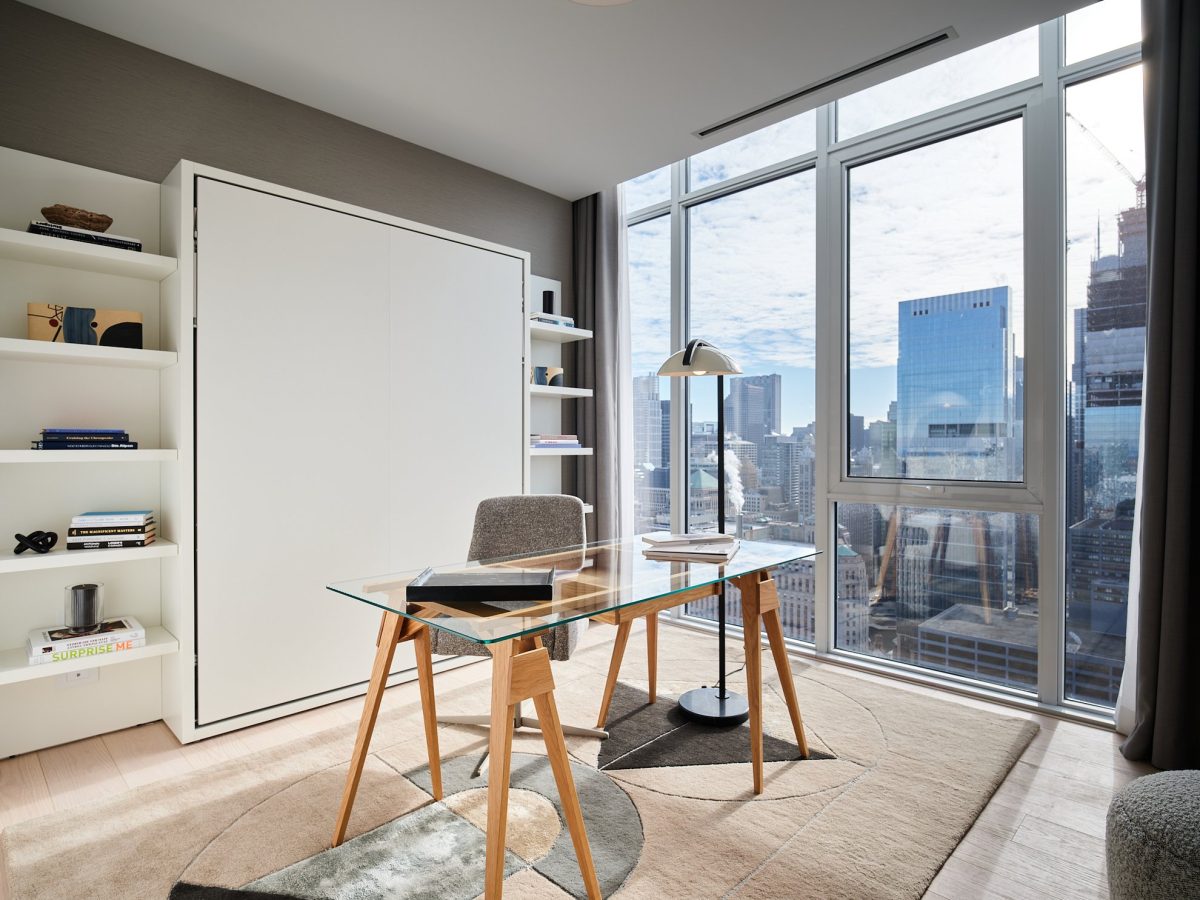A modern home office with a glass desk, gray chair, bookshelf, and a large window offering a city skyline view. Natural light fills the room, which features a neutral color palette and minimalist decor.