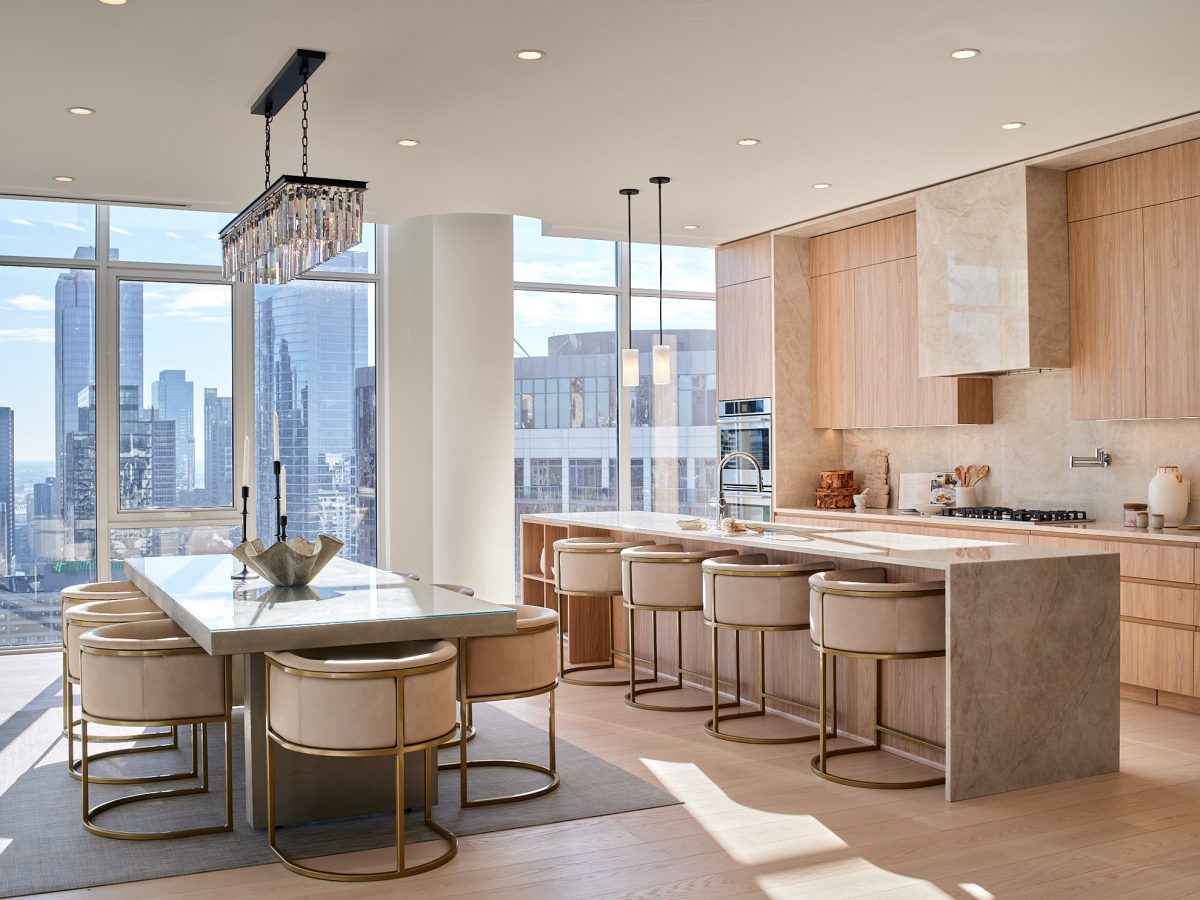 Modern kitchen and dining area with light wood cabinets, marble countertops, beige bar stools with gold accents, a large crystal chandelier, and floor-to-ceiling windows offering a city skyline view.