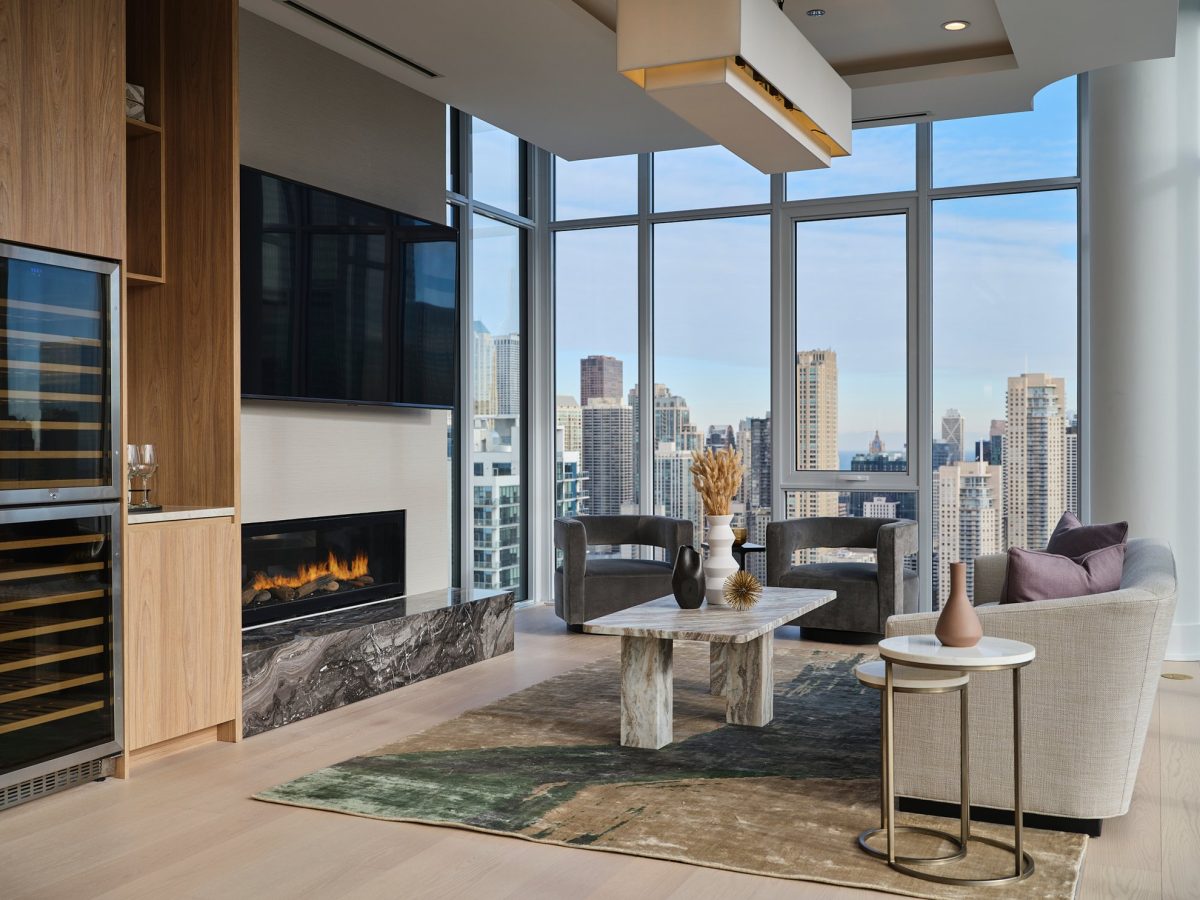 Modern living room with floor-to-ceiling windows showing a city skyline, a marble fireplace, a large TV, neutral-colored furniture, and a marble coffee table on a textured rug. Natural light fills the space.