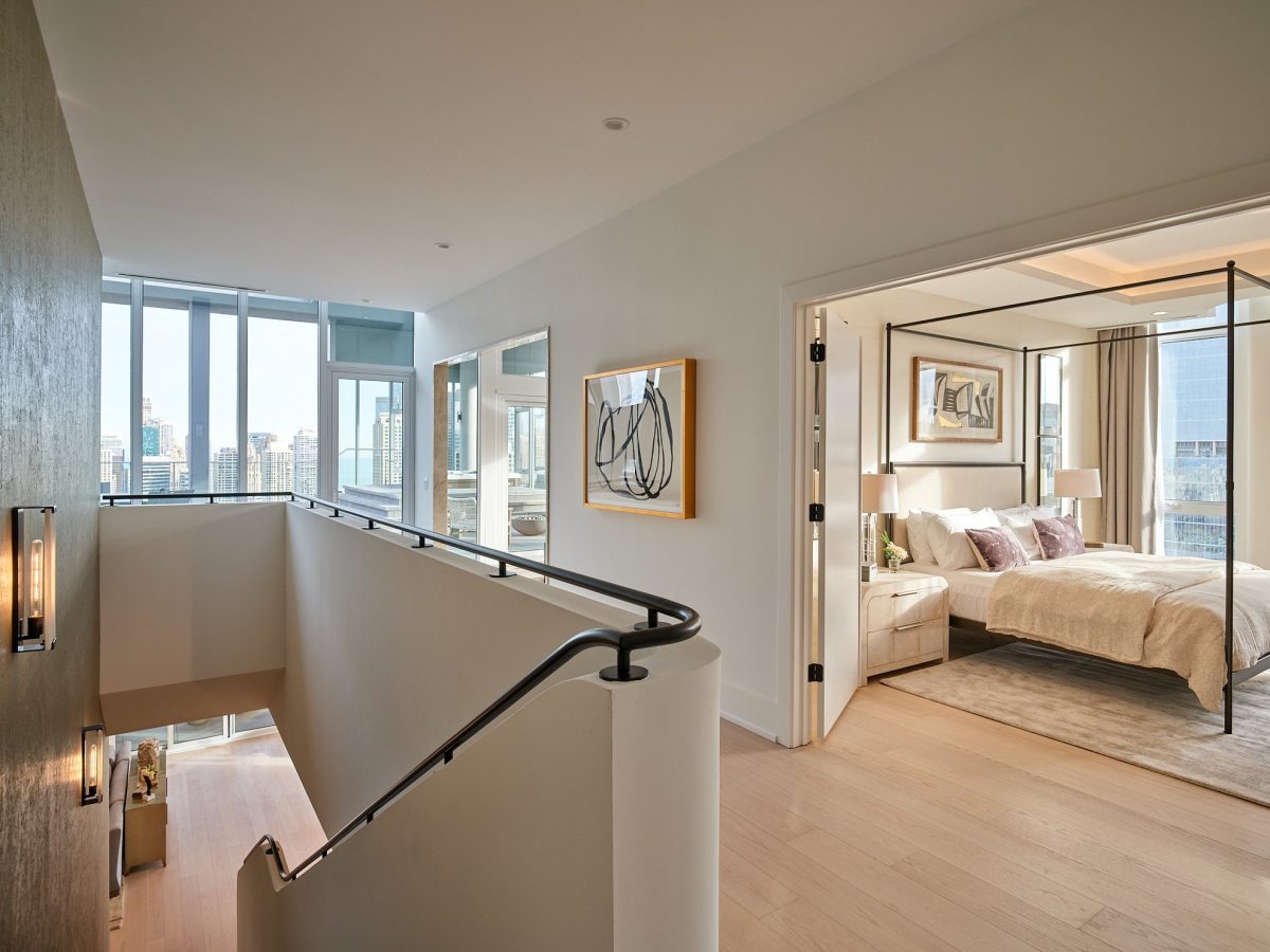 A modern, bright apartment hallway with light wood floors, glass railings, and artwork on the walls, leading to a spacious bedroom with a large bed and city views through floor-to-ceiling windows.
