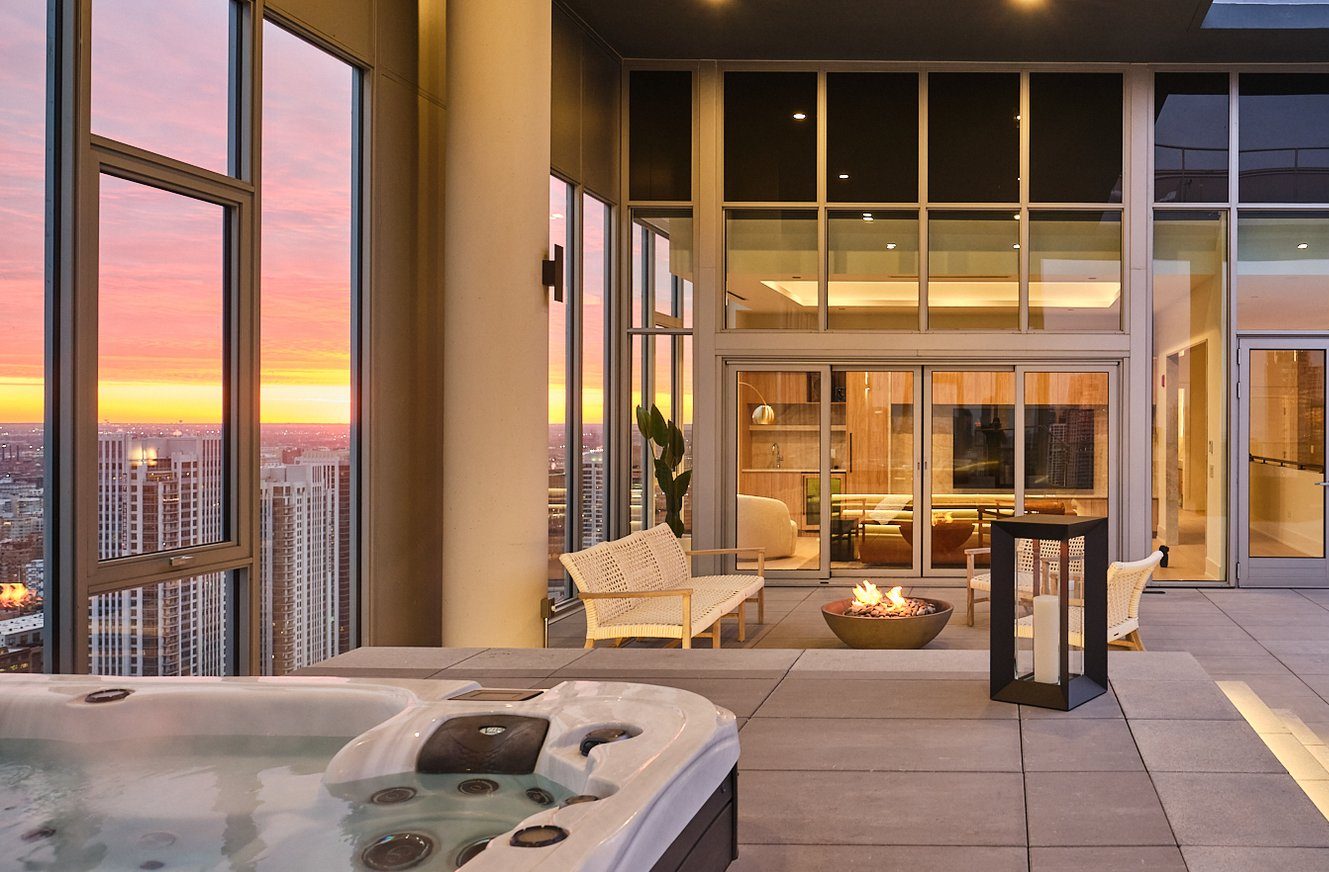 Modern rooftop terrace at sunset featuring a hot tub, white patio chairs around a fire pit, and floor-to-ceiling windows overlooking a city skyline. Warm lighting enhances the cozy, luxurious atmosphere.