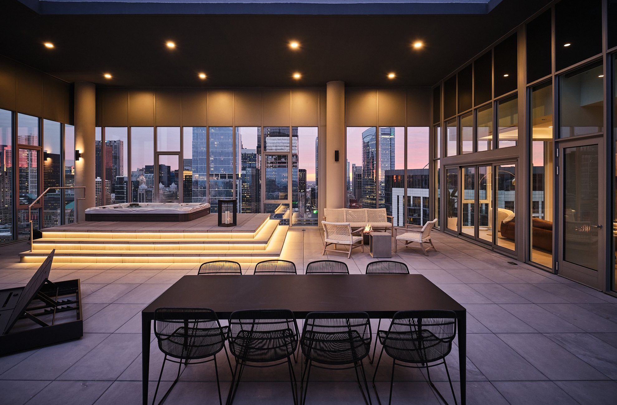 Modern rooftop terrace at sunset with a dining table and chairs in the foreground, a lounge area, and a raised hot tub with glowing steps, surrounded by city skyscrapers and large glass windows.