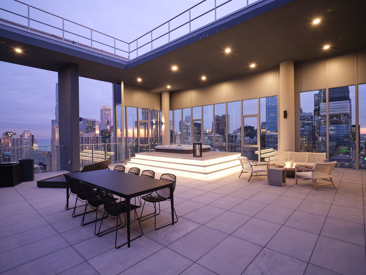 Modern rooftop terrace at dusk with city skyline views, outdoor dining table with chairs, lounge area with armchairs around a fire pit, and a central illuminated platform under a covered section.