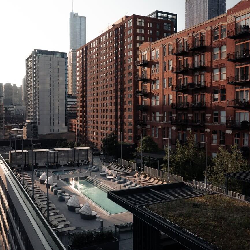 A rooftop swimming pool with lounge chairs and bean bags is surrounded by tall brick and modern high-rise buildings in a city during the day. The area appears calm and empty.