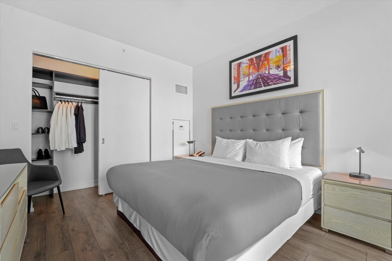 A modern bedroom with a neatly made bed, gray tufted headboard, two bedside tables with lamps, an open closet with clothes and shoes, and a colorful cityscape artwork on the white wall above the bed.