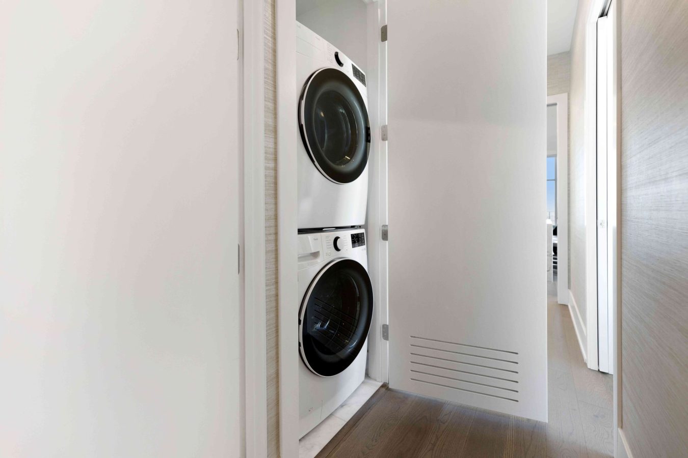 A stacked washer and dryer are tucked into a small closet with a white sliding door, located in a hallway with wooden flooring and light-colored walls.