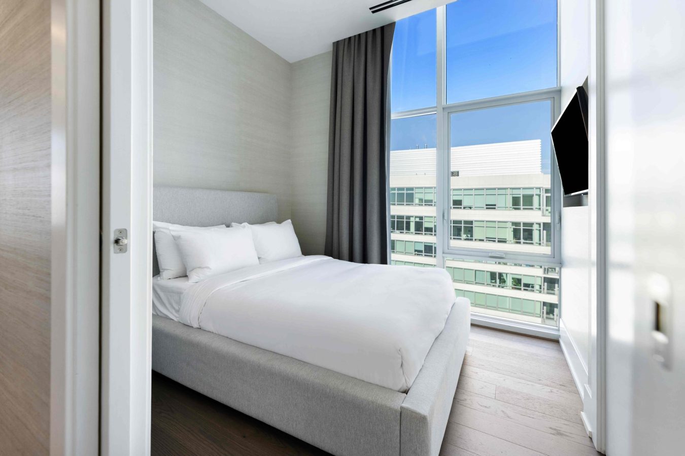 A modern bedroom with a large bed, white bedding, light gray headboard, floor-to-ceiling window with gray curtains, wooden floor, and a wall-mounted TV. Bright daylight streams in from the window.